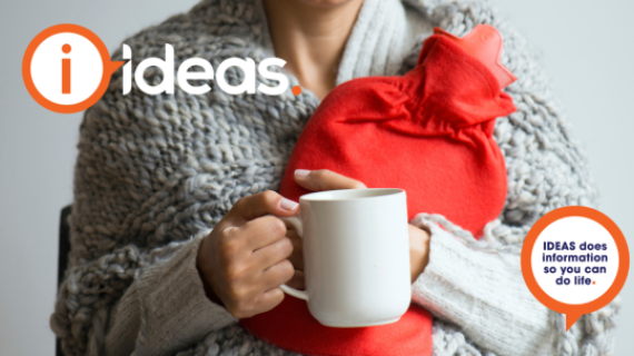 A person wrapped in a chunky grey blanket, holding a cup and hot water bottle in a cover.