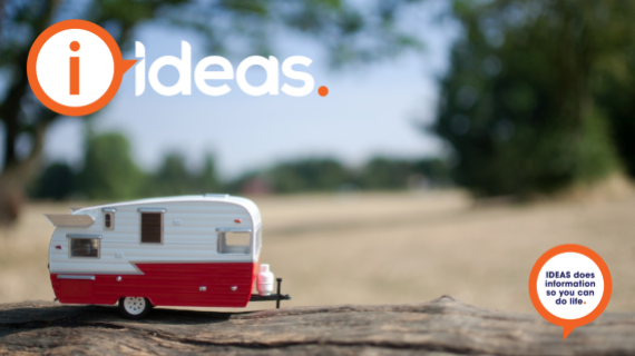 Concept photo of caravaning. A vintage style toy caravan, in red and white, sits on a piece of wood in the outdoors.