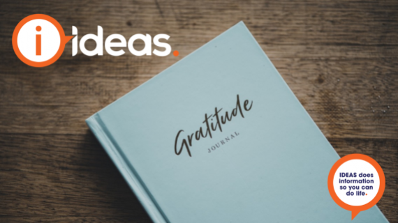 A pale blue hard bound book, with the words "gratitude journal" written on the front cover in a mixture of cursive and plain text rests on a timber tabletop.