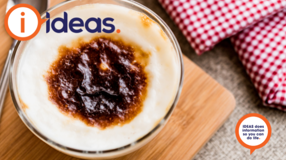 A bowl of rice pudding, with a dark patch on the skin top. ON a bamboo board with a spoon and checked serviette.