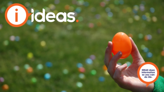 A small hand holds up an orange coloured plastic easter egg. In the background are blurred easter eggs on green.