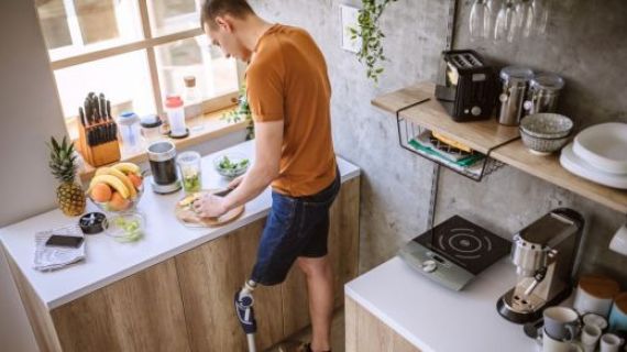 A photograph of a young male with prosthetic leg. He is standing in a kitchen, chopping food. In front of him is a blender to make a smoothie with.
