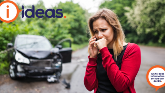 A woman with a worried look on her face and hand clenched to her jaw is calling someone on a smartphone. Behind her is a damaged car.