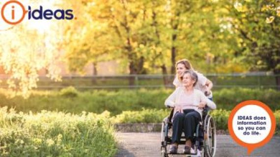 Elderly lady in a wheelchair being pushed by her carer. Ideas logo in the image.