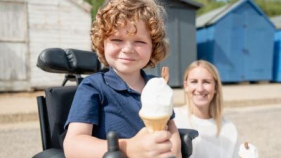 Young boy in wheelchair holding an ice-cream. Beside him is a woman holding an ice-cream. In the background are beach huts.