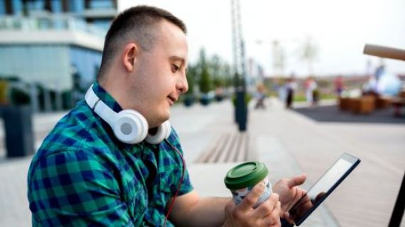A teen male living with intellectual disabilty, sitting looking at a tablet device, He is holding a reusable takeaway cup and has headphones resting on his neck.of