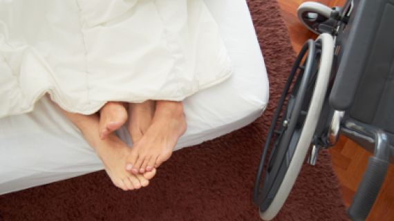 Image of a couple's feet sticking out from the covers in bed and a wheelchair parked alongside.