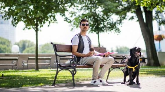 An image of a young man sitting on a park bench. He wears sunglasses, is holding a cane, and beside him is a guide dog.