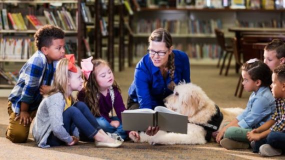 children, teacher and assistance dog sitting on the floor in a library reading a book as a group
