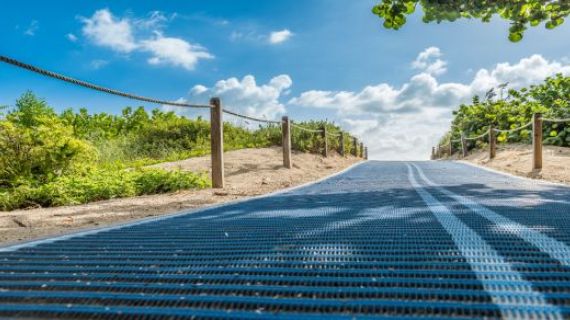 A photograph of a beach pathway with accessible matting. In the background is a blue sky with clouds.