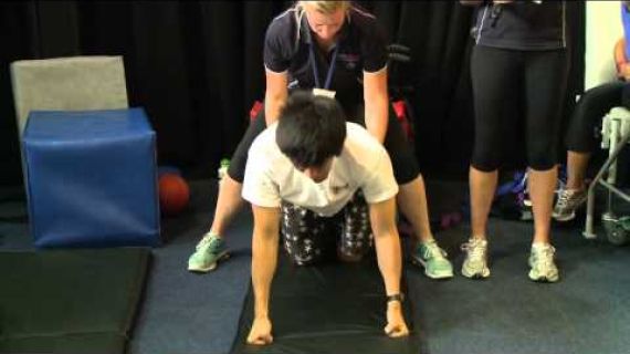 Person on hands and knees with someone behind helping with rehabilitation
