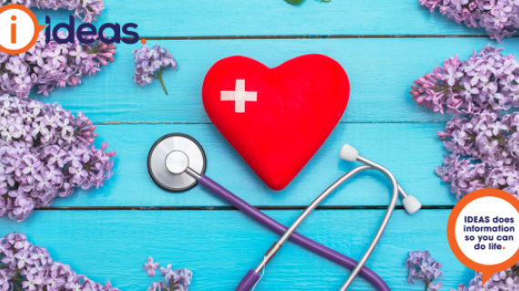 Red heart with bandaid in a cross shape, sit on a blue background. It is surrounded by lilac blooms, and a stethoscope.
