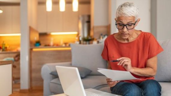 A woman wearing glasses and a red shirt, holds a pen and is checking some paperwork. In front of her is a white laptop.