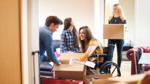 A group of University students are moving into a shared flat. They are surrounded by boxes. One student is in a manual wheelchair.