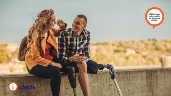Two individuals with lower leg amputations sit on a wall, smiling warmly at each other.