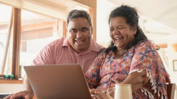 A mature Aboriginal couple sit at a table. In front of them is a laptop, they are both smiling/ laughing at something on the screen. A cup of sits to the left of the woman.