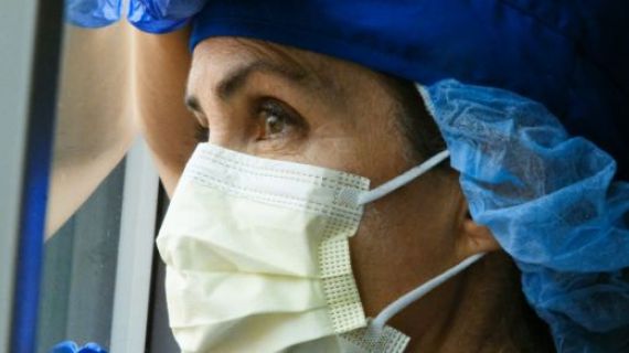 A health worker in PPE resting her hand and head against a window and looking outside.