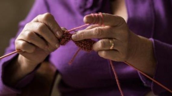 detail of older woman's hands knitting. she is wearing a purple jacket.