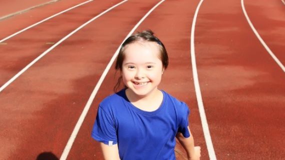 A photo of a young girl with down syndrome smiling at the camera. She is standing on an athletics track.
