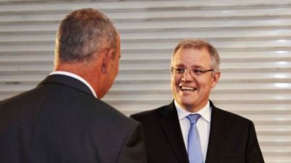 A man in suit and blue tie with glasses smiling and speaking to another man in suit with back facing camera