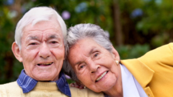 One elderly female is resting their head on the shoulder of another elderly man.