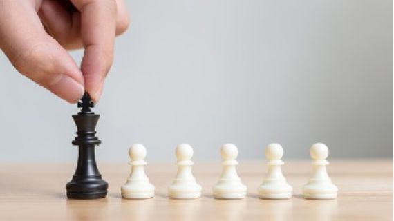 Image of hand holding black chess piece (King) with a row of white chess pieces (pawns)