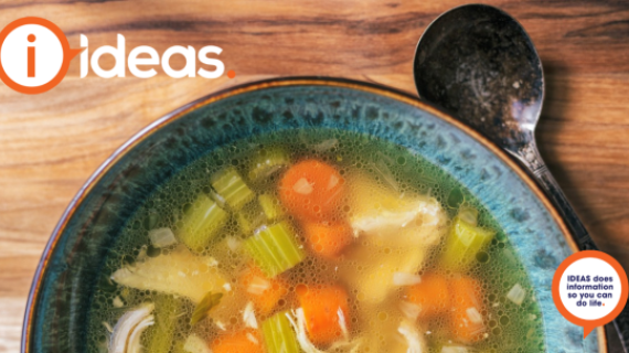 A blue bowl with a brown edge sits on a timber table. Beside it is a spoon. In the bowl is broth, celery, chicken, carrot and onion. A bowl of Chicken soup.