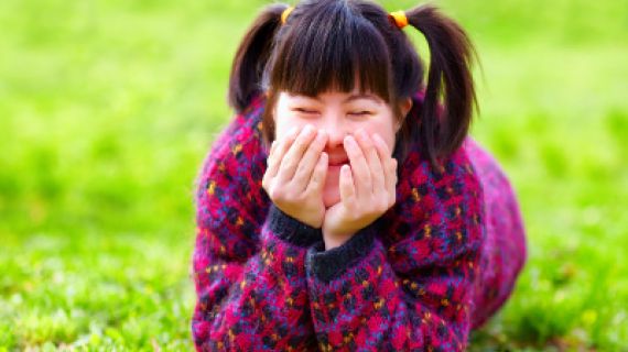 A young girl with disability is laying in the grass, she has her eyes closed, is smiling with her hands partly covering her face.