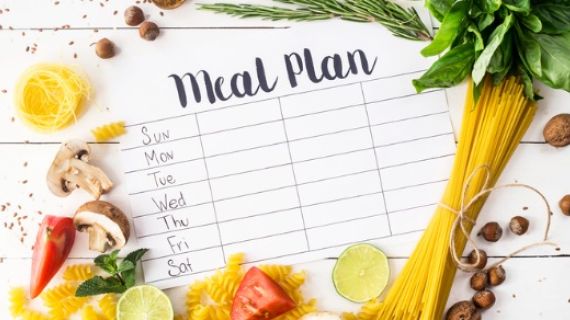 A printed piece of paper with the heading "Meal Plan" has days of the week and columns to place meals. The paper is surrounded by foods like pasta, herbs, vegetables, fruits, nut and spices,