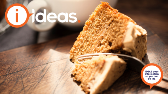 A slice of coffee cake rest on a board, a fork is cutting into the cake. A white cup is in the background.