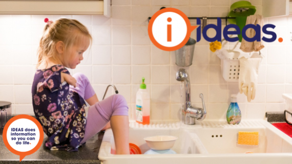 A young girl sits on the kitchen bench beside the sink. She washes the dishes with her feet.