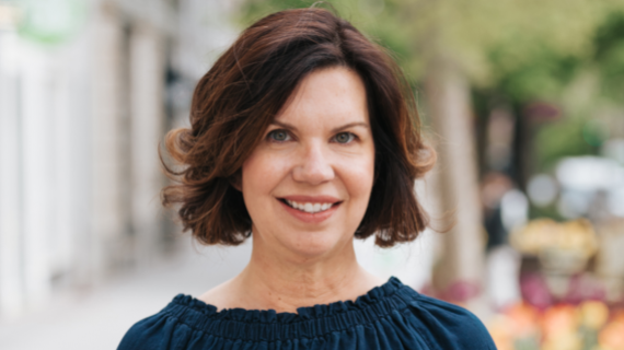 A portrait image of a woman with brown hair and a blue top, she is smiling and looking a the camera. She has an invisible disability.