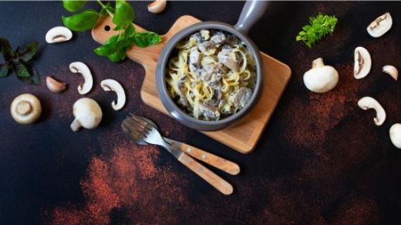 A concept photo of mushroom stroganoff, a dish with mushrooms and pasta, aongside is a knife and spoon. Around the bowl are mushrooms, basil, herbs and paprika.