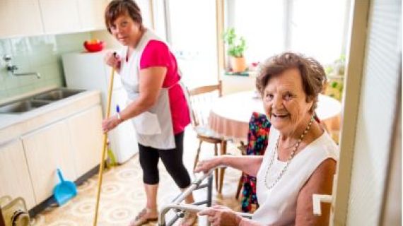 A senior lady sitting and a care giver sweeping the floor.