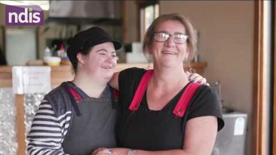 Victoria standing with a colleague arm in arm in the cafe that they work in.