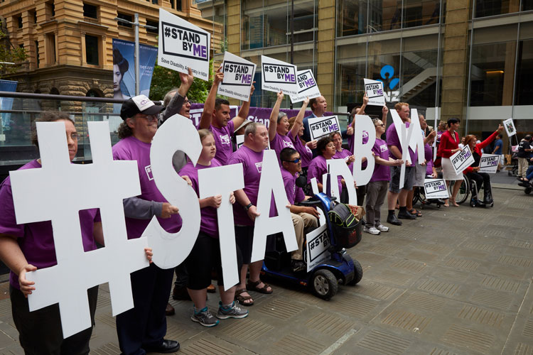 A group of women and children and two men dressed in a mix of purple and white clothing are standing outside a city building, holding protest signs 