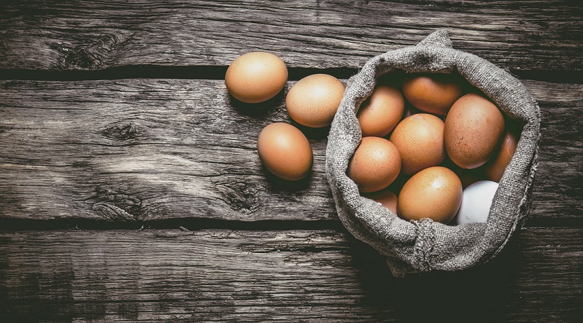 Eggs lying in a basket on a wooden table with some spilled out beside the basket.