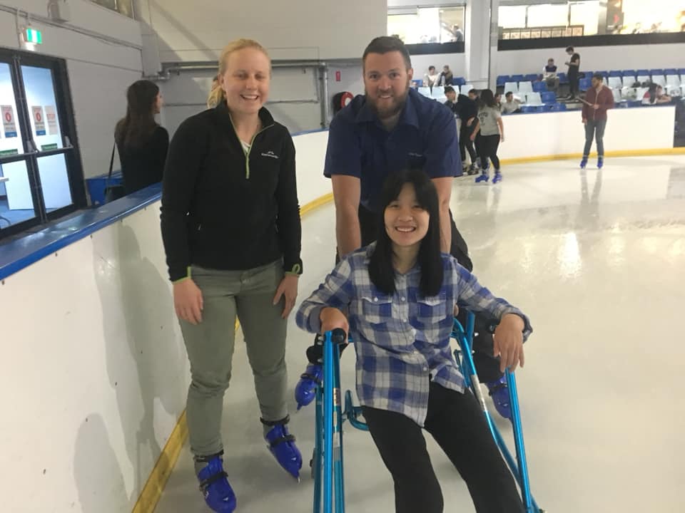 Nicole and two friends standing on an ice skating rink. Nicole is supported and in a semi-seated position by a mobility aid, all are smiling broadly.